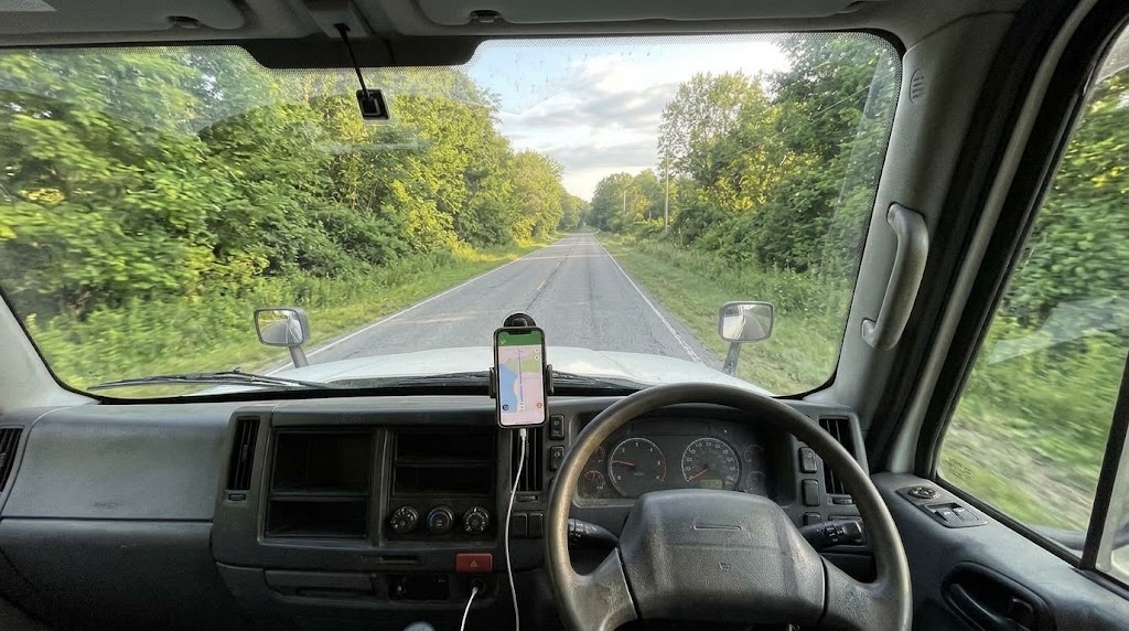 初夏の緑の道を走るトラックの運転席。ダッシュボードのホルダーにスマートフォンが固定されている。